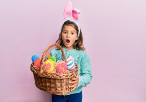 Little beautiful girl wearing cute easter bunny ears holding wicker basket with colored eggs celebrating crazy and amazed for success with open eyes screaming excited.