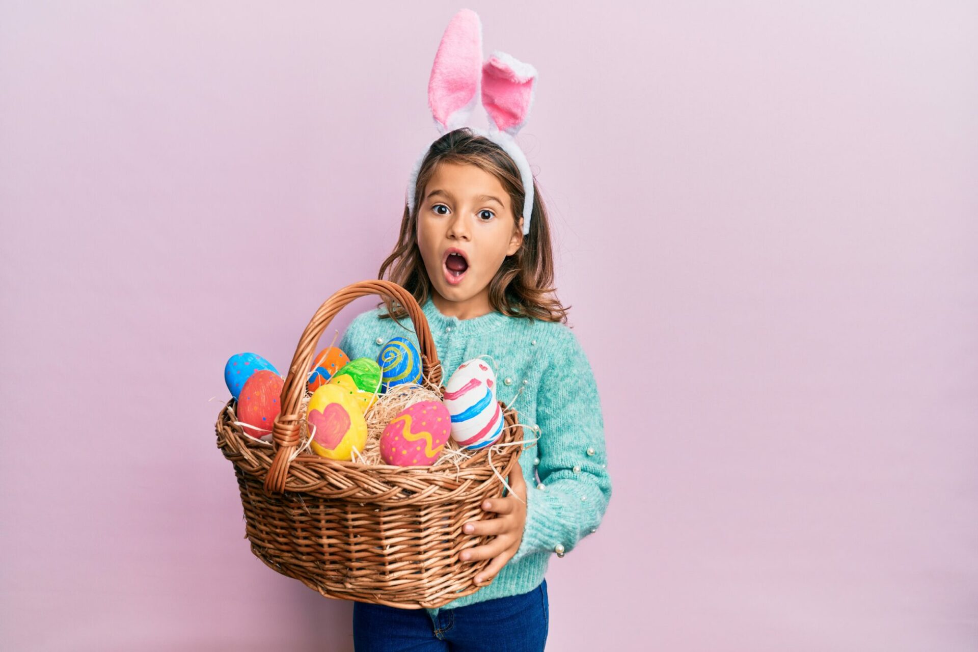 Little beautiful girl wearing cute easter bunny ears holding wicker basket with colored eggs celebrating crazy and amazed for success with open eyes screaming excited.